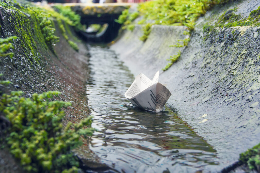 A small paper boat floating in a shallow, concrete drainage channel, surrounded by green moss and plants