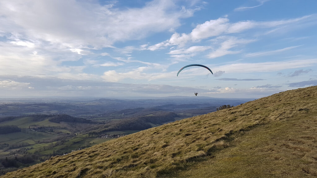 A paraglider floating in the air over a green hill on a cloudy day with blue sky with a background view of rolling hills